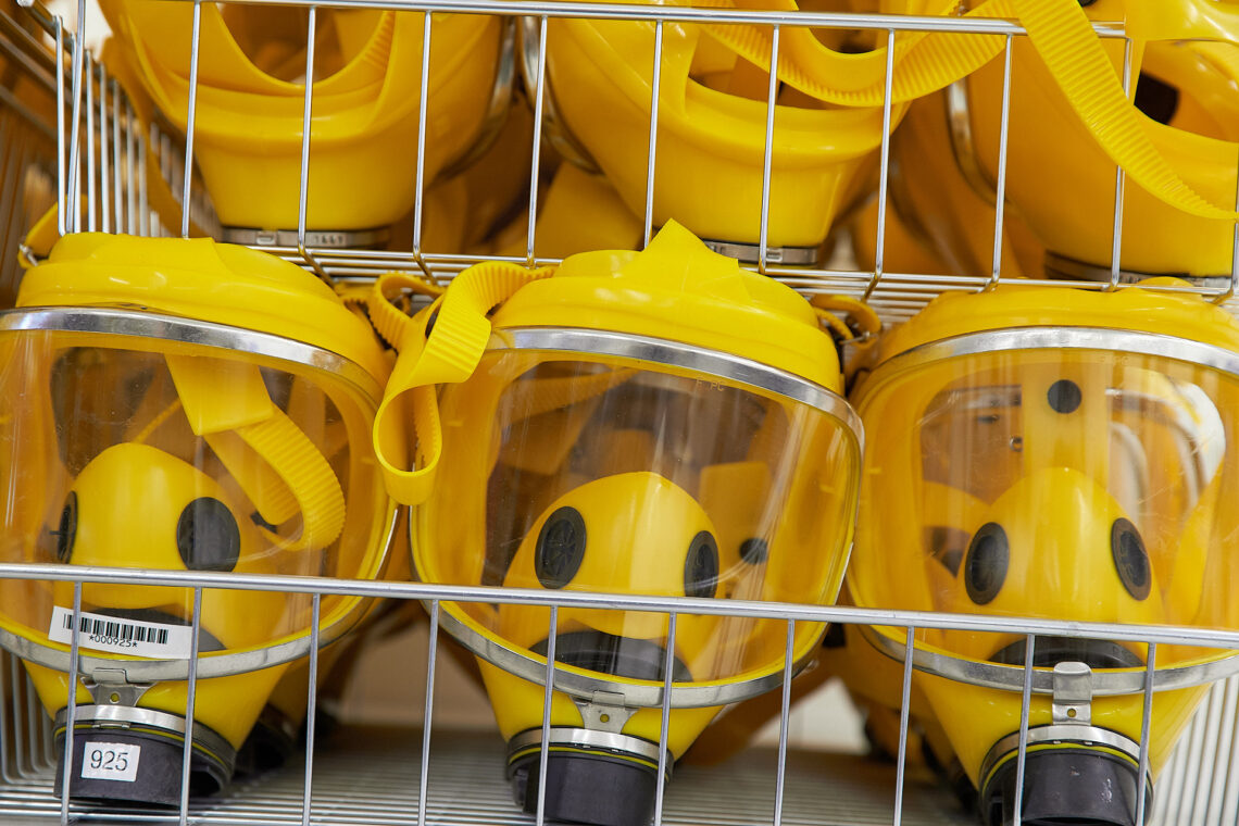 Nahaufnahme mehrerer gelber Atemschutzmasken mit durchsichtiger Sichtscheibe, die dicht an dicht in einem Metallgitterkorb liegen. Die Masken sind mit gelben Kopfbändern versehen.