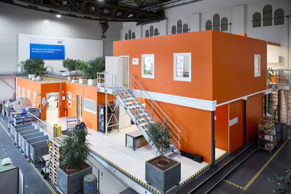 Zweistöckiges, orangefarbenes Modulgebäude im Inneren einer großen Werkshalle mit Fenstern, einer außenliegenden Metalltreppe und Kübelpflanzen auf beiden Ebenen. An der Hallenwand hängt ein Banner mit der Aufschrift 'BTI – Servicepartner für Arbeitssicherheit, Brandschutz und Gebäude'. Im Erdgeschoss geht ein Mitarbeiter der Firma BTI zwischen grauen Rollcontainern und Paletten, rechts stehen weitere Gitterwagen mit Kartons.