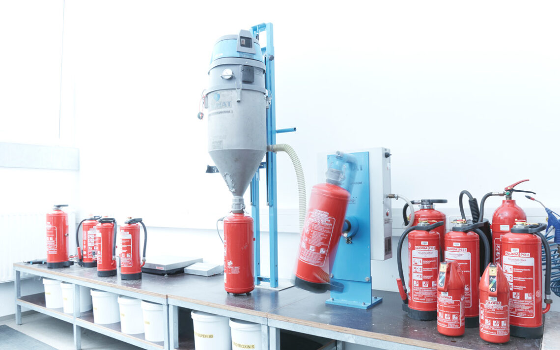 Arbeitsplatz zur Befüllung und Wartung verschiedener roter Feuerlöscher mit technischen Geräten und mehreren Löschern auf einer Werkbank.