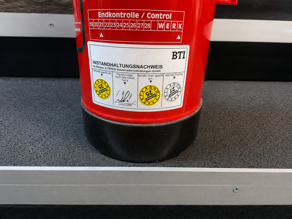 Nahaufnahme eines roten BTI-Feuerlöschers mit Prüf- und Instandhaltungsnachweis auf einem dunklen Metallregal.