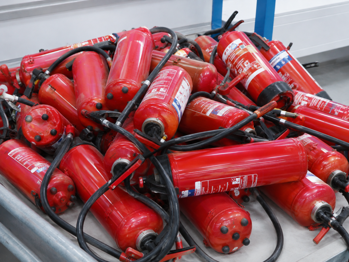 Mehrere ausrangierte rote Feuerlöscher mit Schläuchen und Griffen in einem hellen Industriebereich.