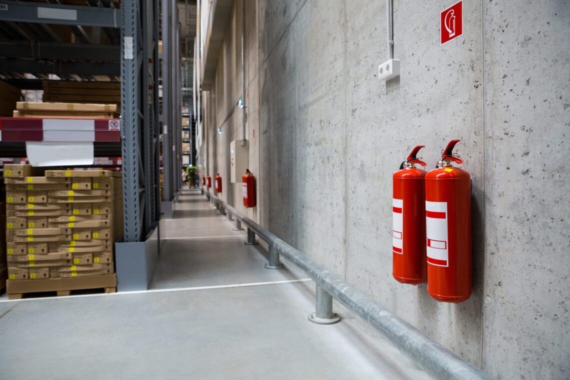 Zwei rote Feuerlöscher an einer Betonwand in einer Industrie- und Lagerhalle mit Regalen und Paletten im Hintergrund.